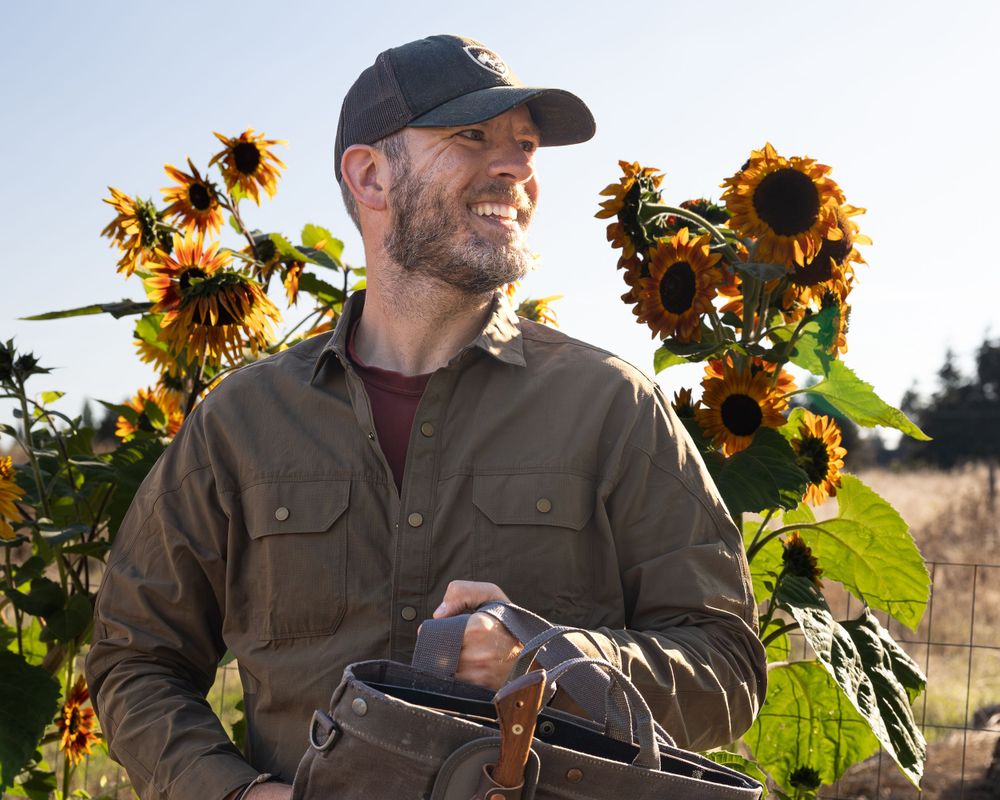 Person in work jacket stands among blooming sunflowers in late afternoon light.