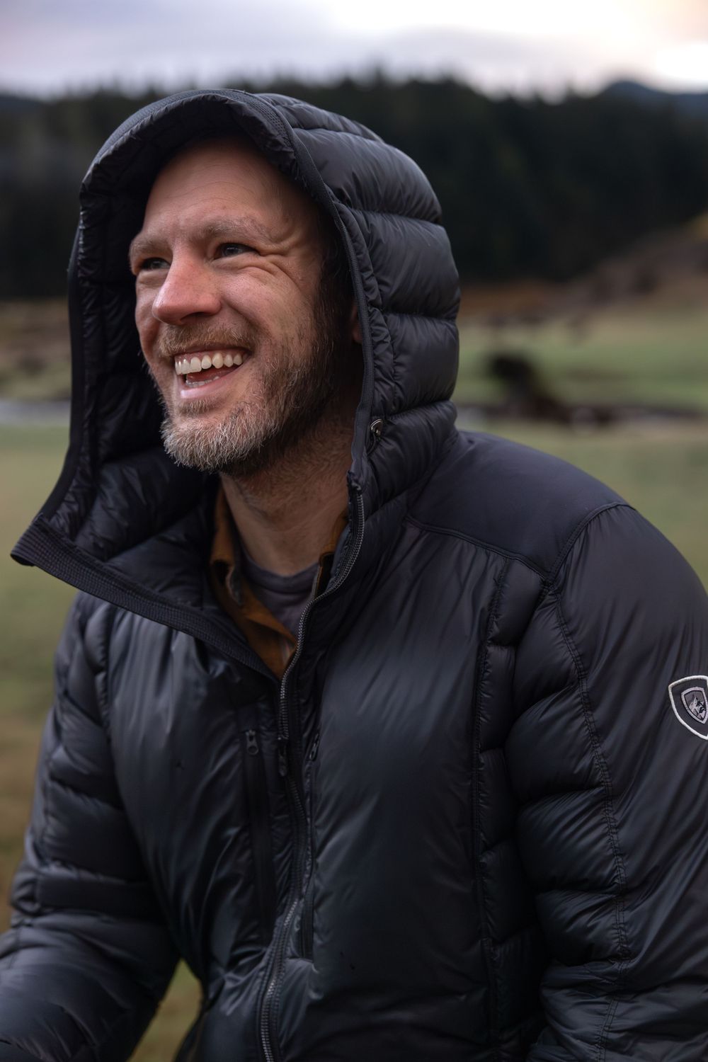 Person in black puffy winter coat smiling outdoors in nature.