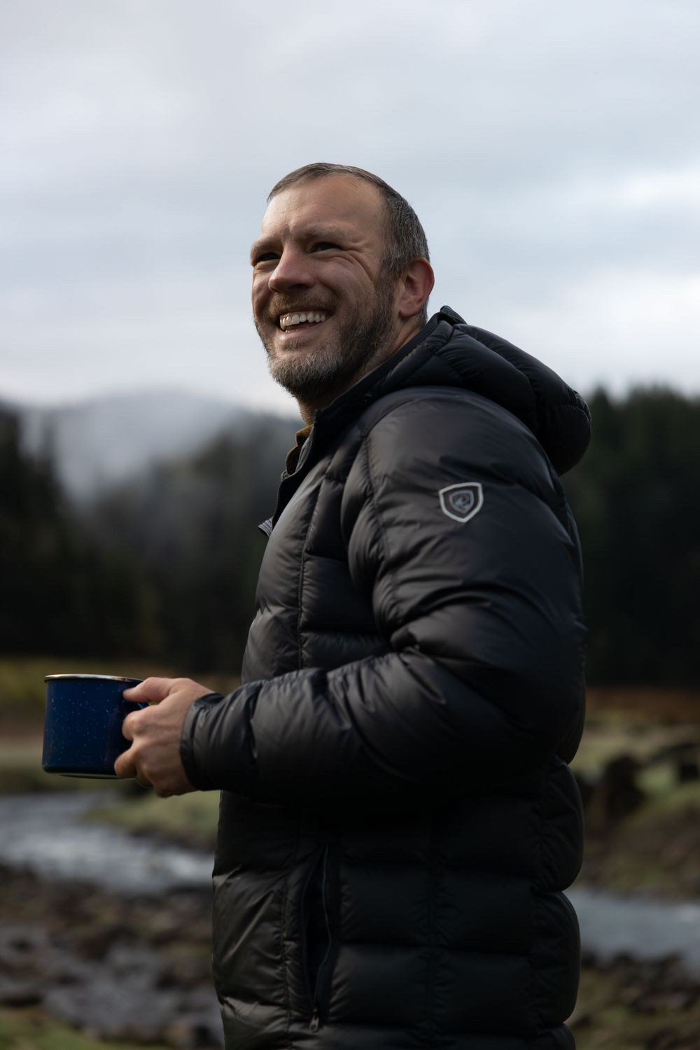 Person wearing black winter coat holding blue mug outdoors in nature.
