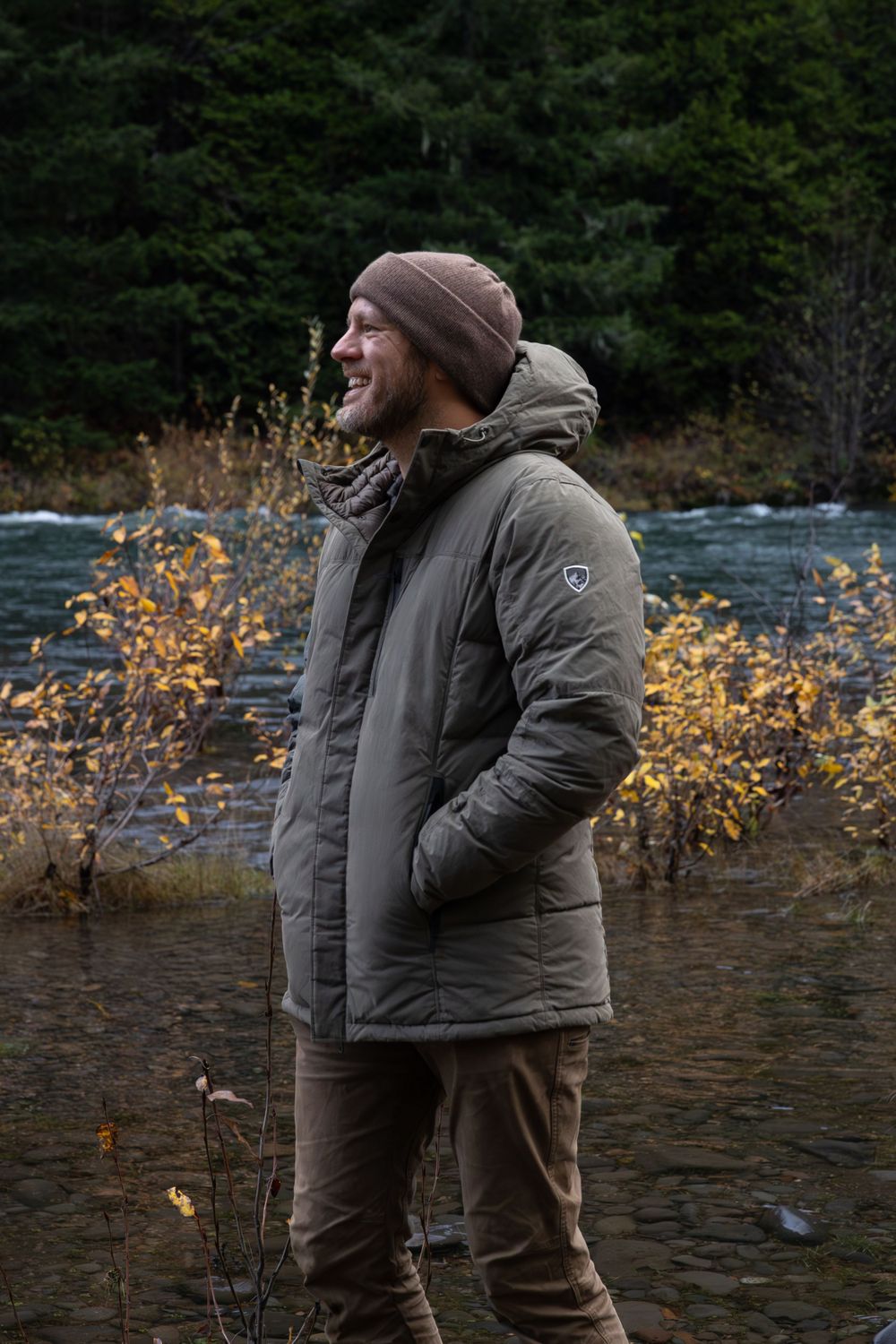 Person in warm winter attire stands near a river with autumn foliage in the background.