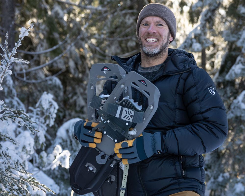 Person holding snowshoes smiles while enjoying a snow-covered mountain environment.
