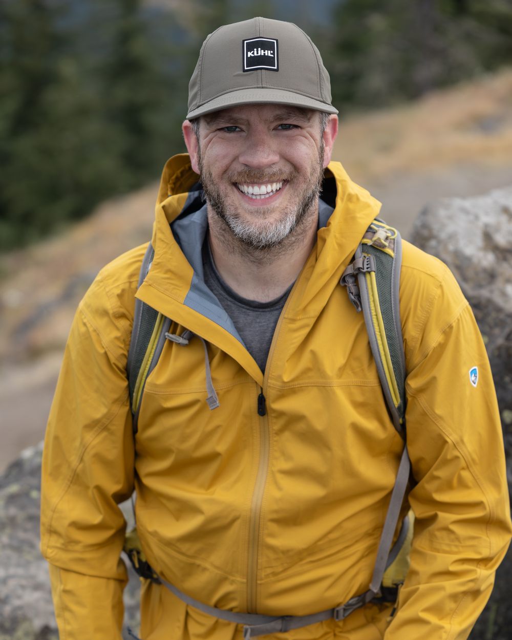 Outdoor enthusiast wearing yellow rain jacket and gray cap against mountain backdrop.