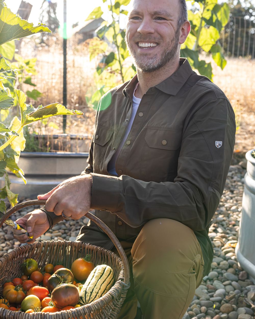 Person in brown jacket harvesting fresh vegetables from garden basket in sunlight.