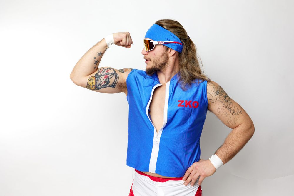Athletic figure in blue workout vest and sunglasses flexes muscles while posing against white background.