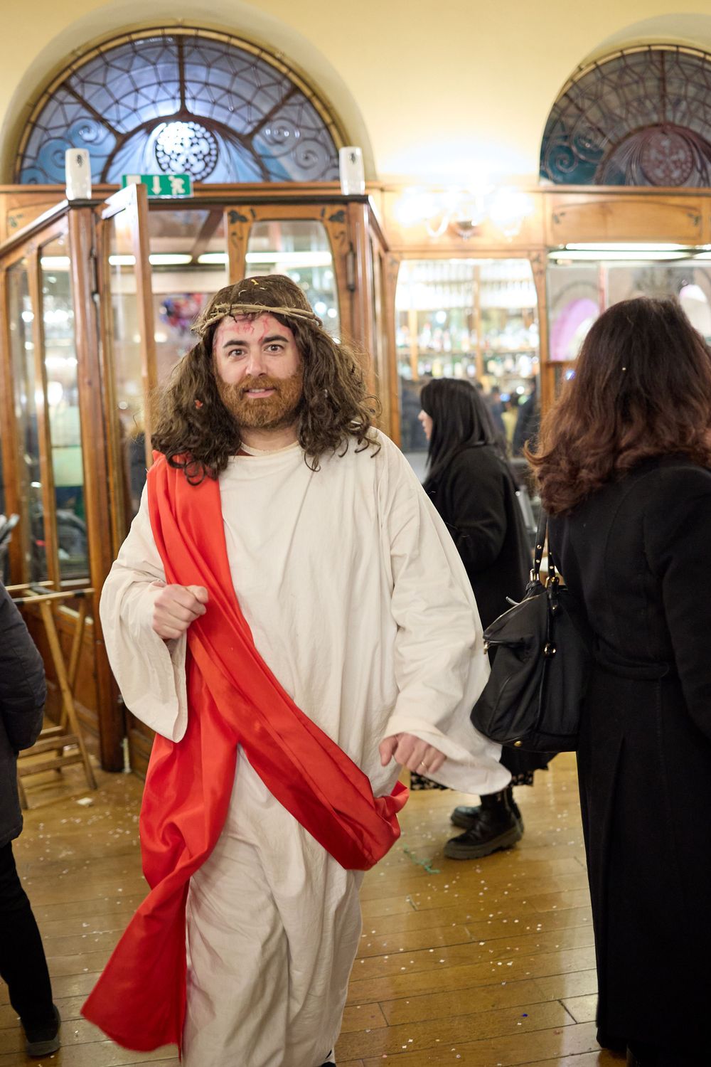 Person in white robe with red sash and crown of thorns stands in historic building with arched windows.