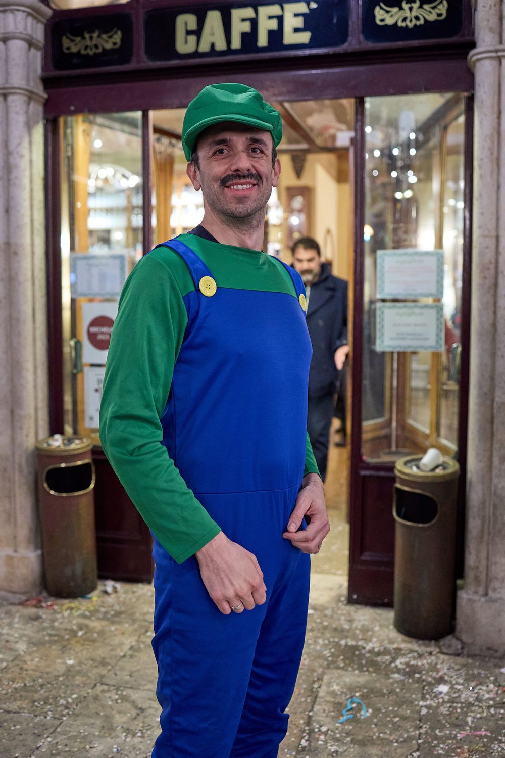 Someone dressed in a classic green and blue video game plumber costume poses outside a cafe storefront.