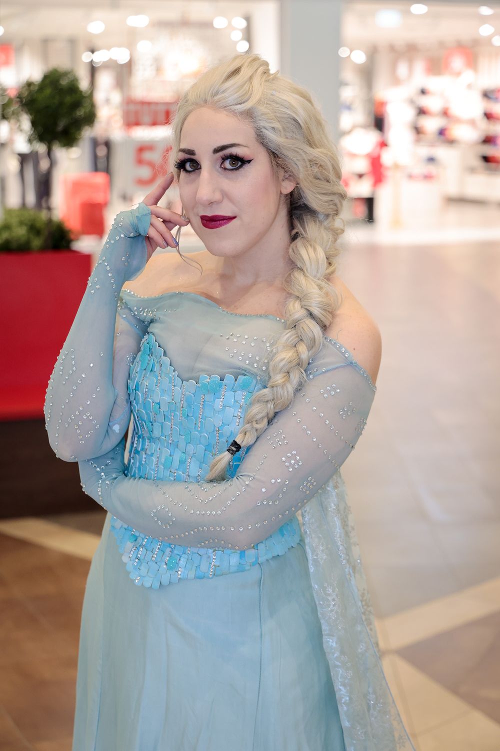 A cosplayer wearing an ice blue sparkly dress and long braid poses in a shopping mall setting.