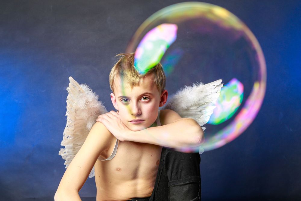 A young person wearing angel wings poses against a blue background with an iridescent soap bubble floating nearby.
