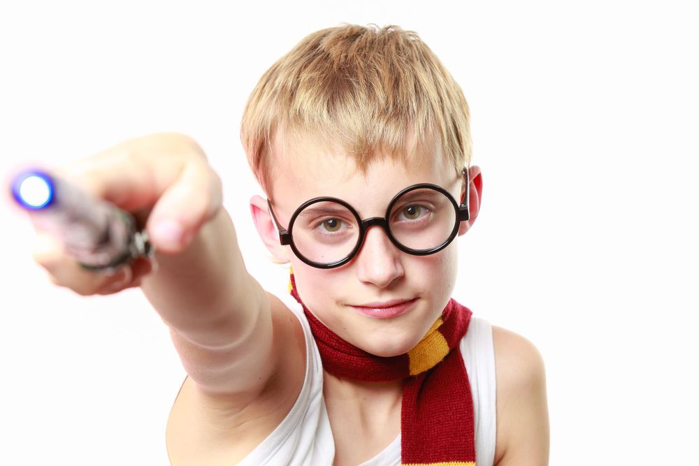 A person wearing round glasses and a red scarf points a wand forward while cosplaying as a wizard.