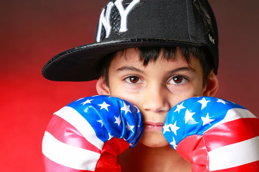 Young sports fan wearing New York Yankees cap poses with American flag boxing gloves against red background.
