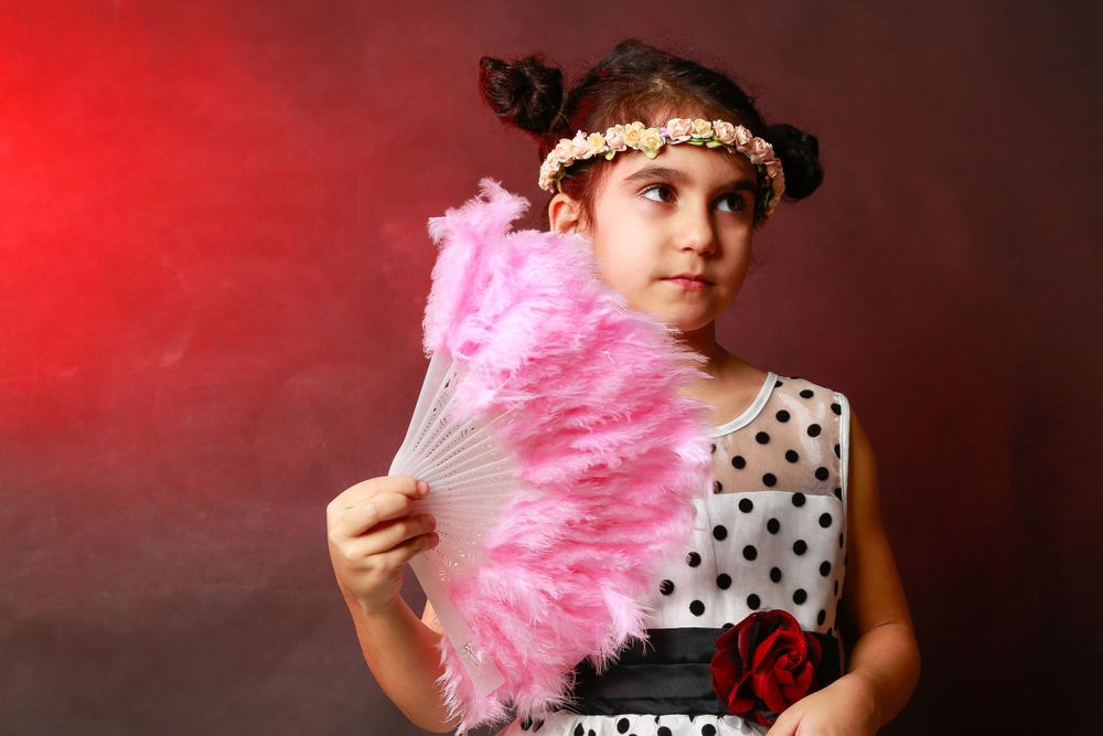 A dancer in polka dot dress holds a pink feathered fan against a dramatic red backdrop.