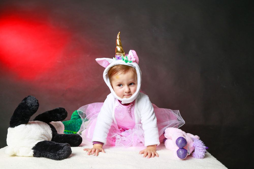 A small child in a pink unicorn costume crawls on a white surface with plush toys scattered around.