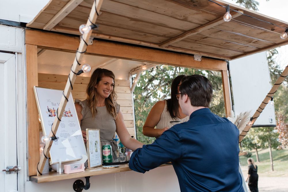 Two people interact at an outdoor wooden service window or food truck counter on a sunny day.