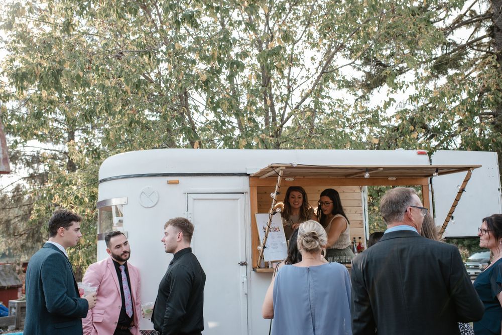 Guests gather around a vintage white camper trailer at an outdoor event while people smile from inside the doorway.