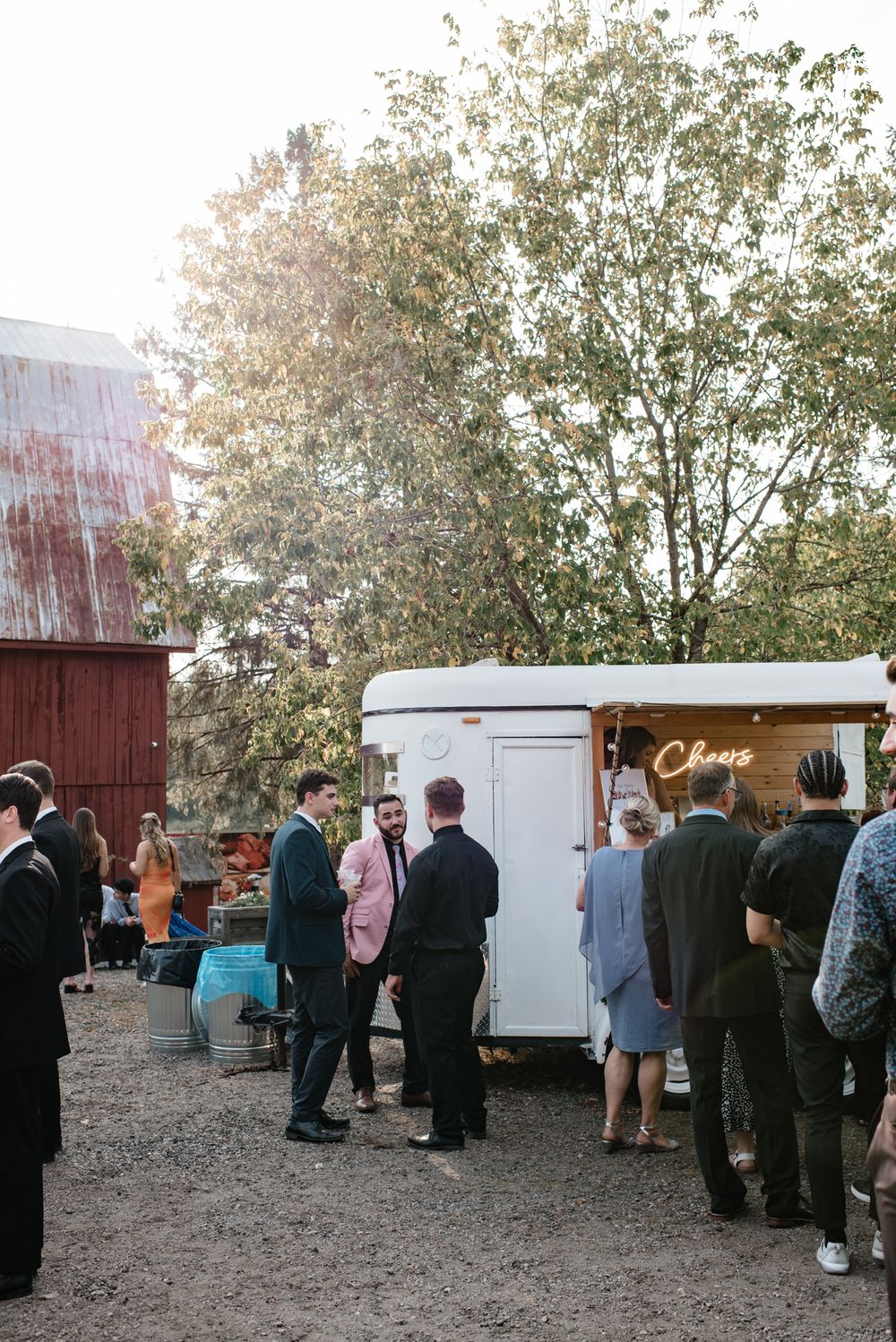 People waiting in line at a white food trailer parked next to a red barn with trees in the background at dusk.