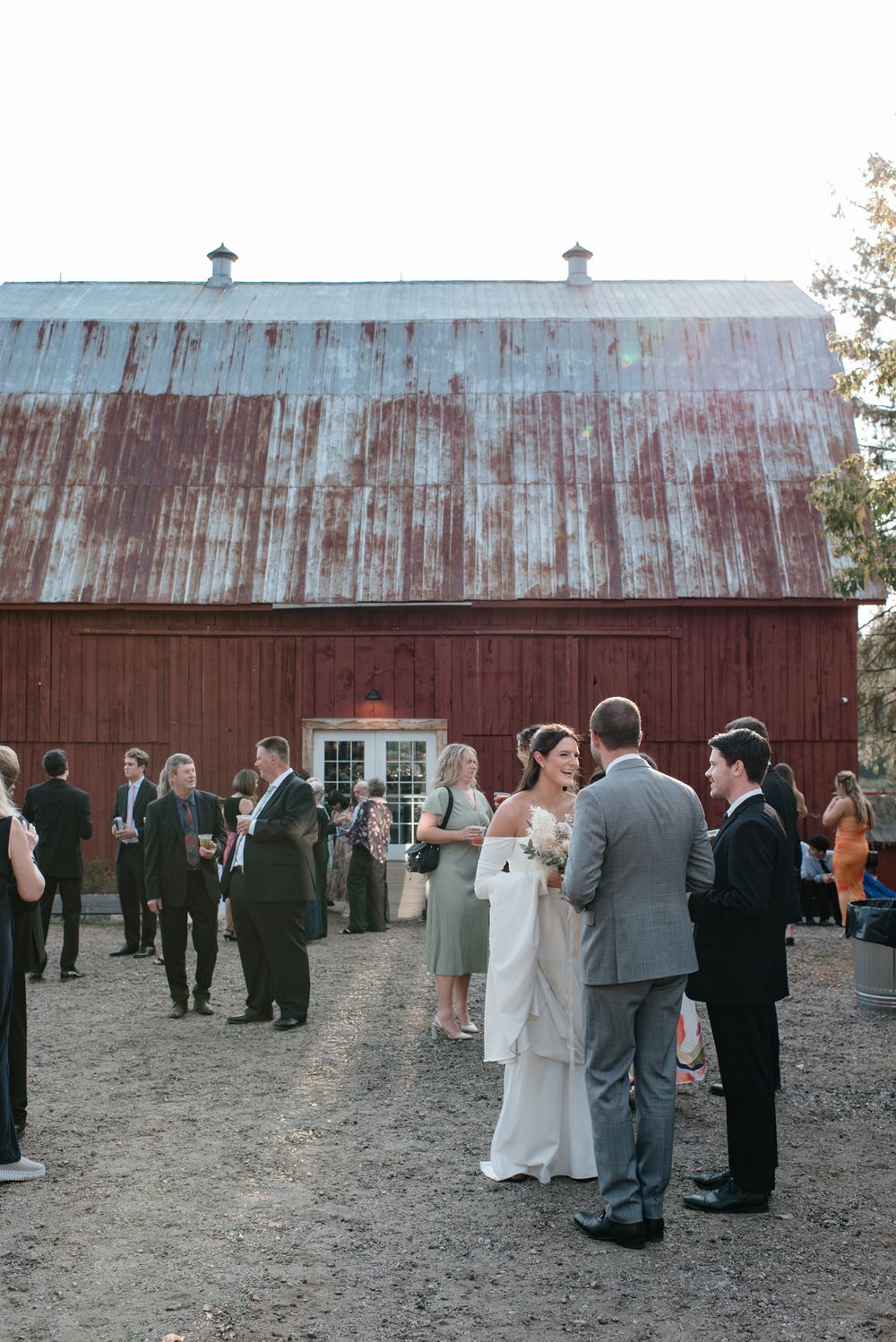 Wedding guests mingle outside a rustic red barn with a weathered metal roof during an outdoor reception.