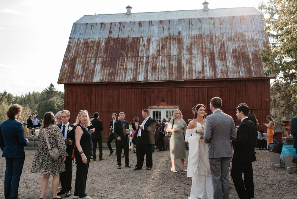 Wedding guests gather outside a rustic red barn during an outdoor reception celebration in a rural setting.