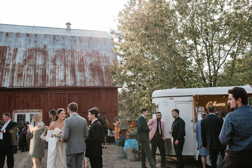 Wedding guests gather for an outdoor reception by a rustic red barn with a food truck parked nearby.