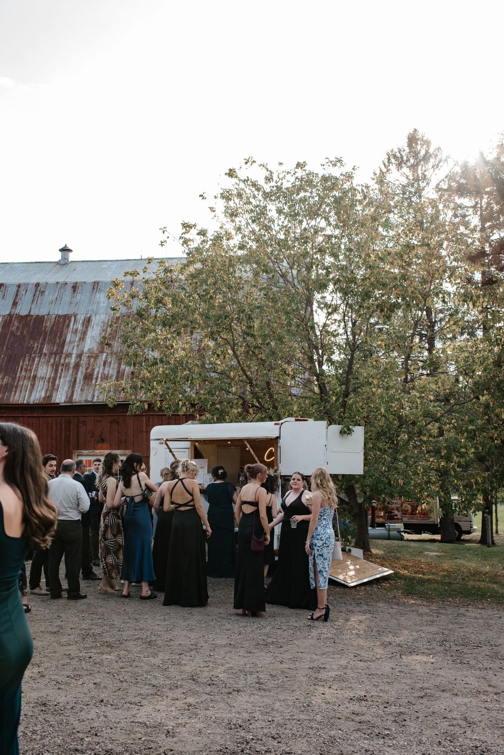 Wedding guests gather outside a rustic barn venue with a white mobile bar trailer and flowering trees in the background.