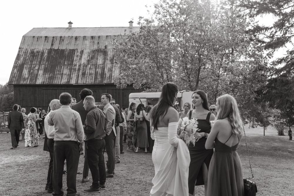 Black and white photo of wedding guests mingling outside a rustic barn during reception.