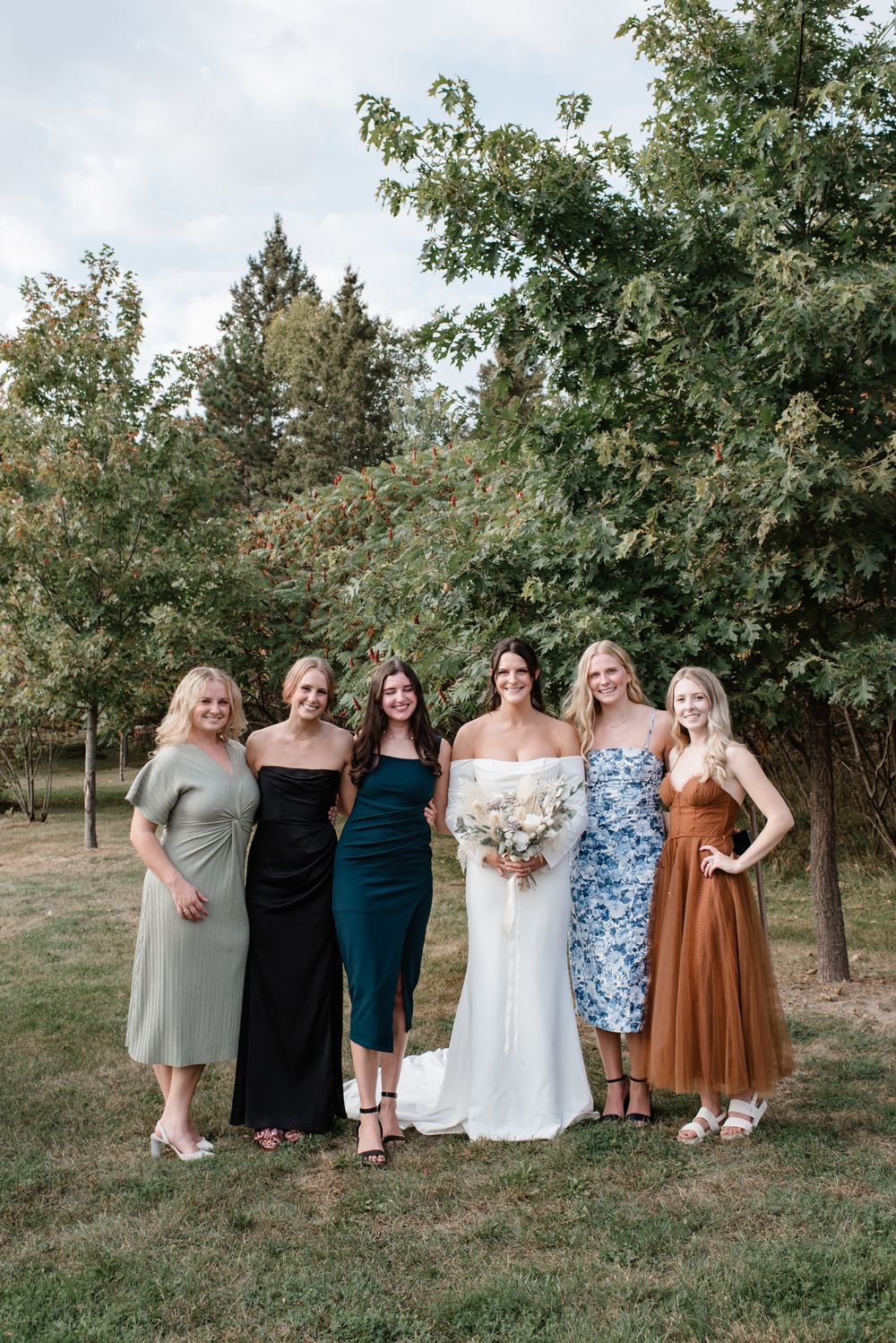 A group of six women in mismatched bridesmaid dresses pose together beneath trees at an outdoor wedding.