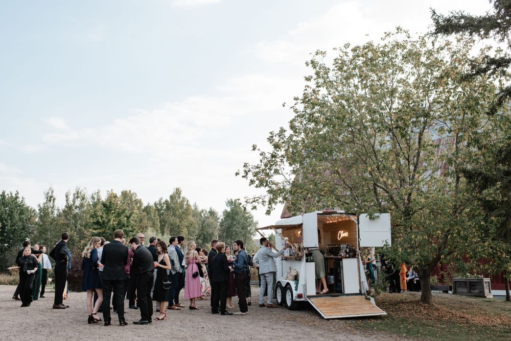 Wedding guests gather around a small wooden trailer photo booth at an outdoor venue surrounded by trees.