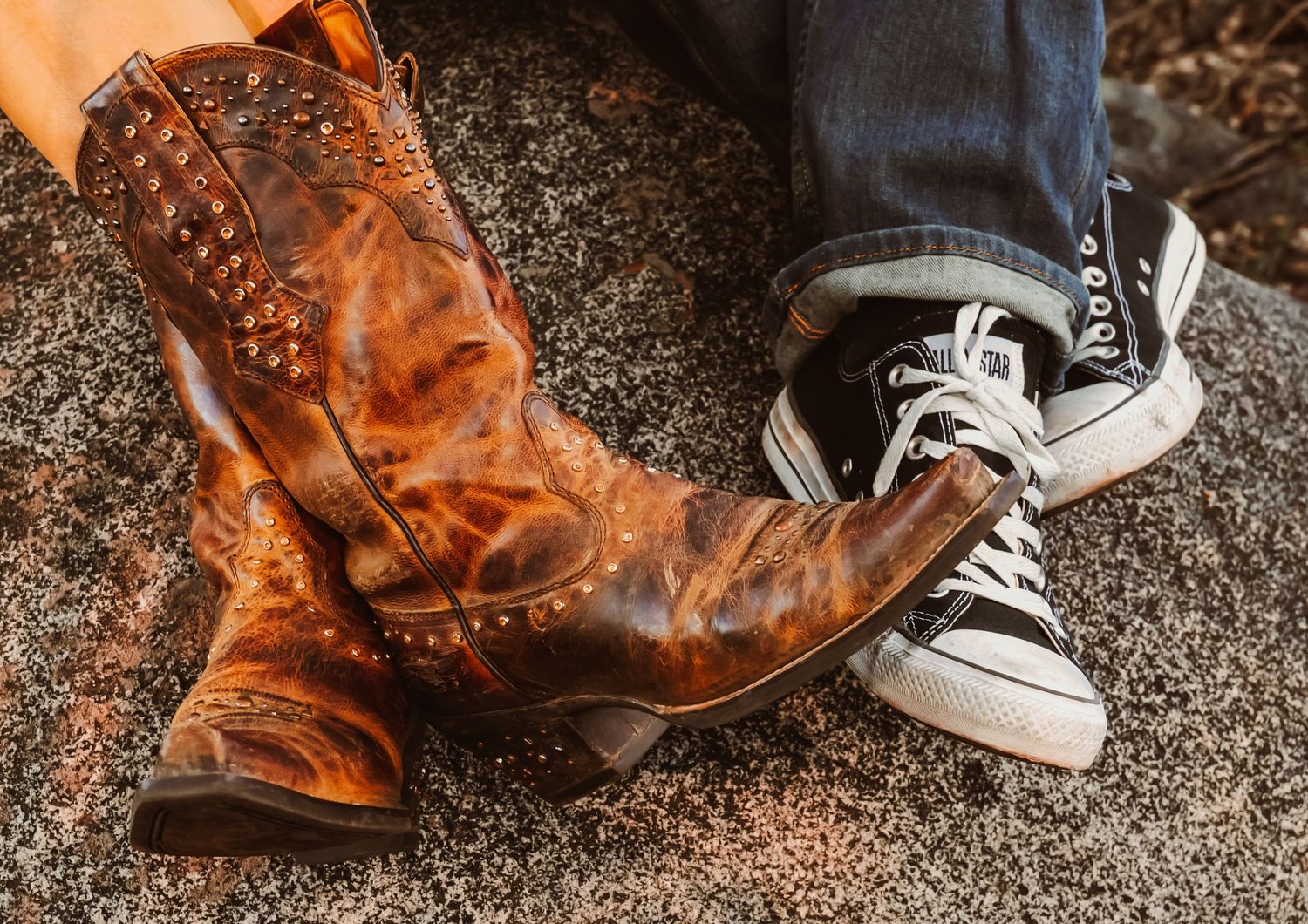 A pair of worn brown leather cowboy boots next to gray canvas sneakers on a concrete surface.