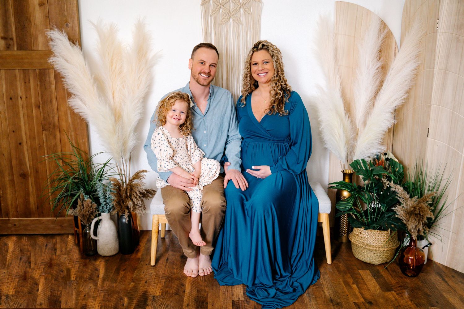 Expecting mother in a beautiful blue gown, sitting in a boho studio holding her belly during a photoshoot with her husband and daughter.