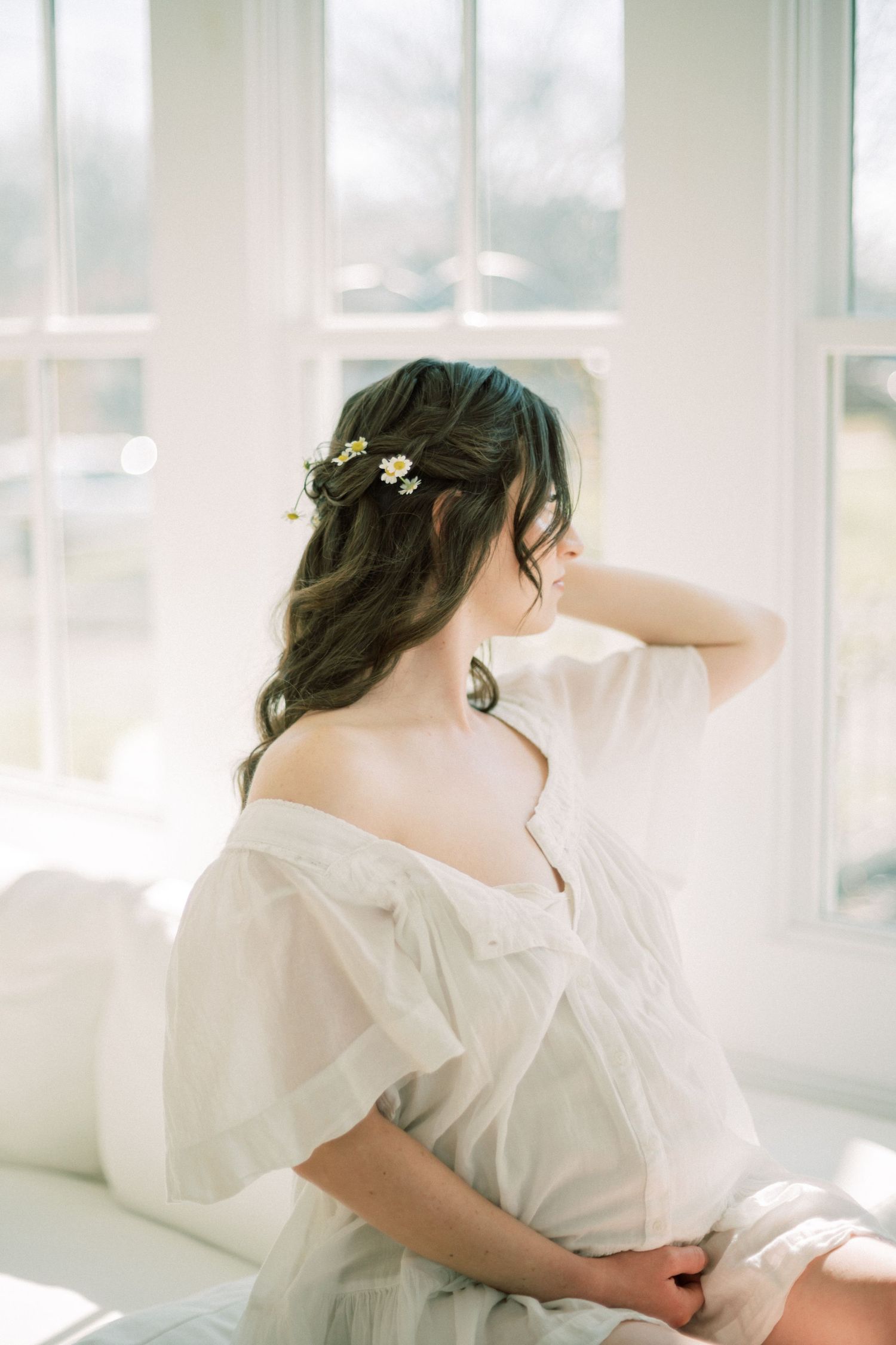 A dreamy ethereal portrait of someone in a white flowing dress with delicate flowers in their hair by a bright window.