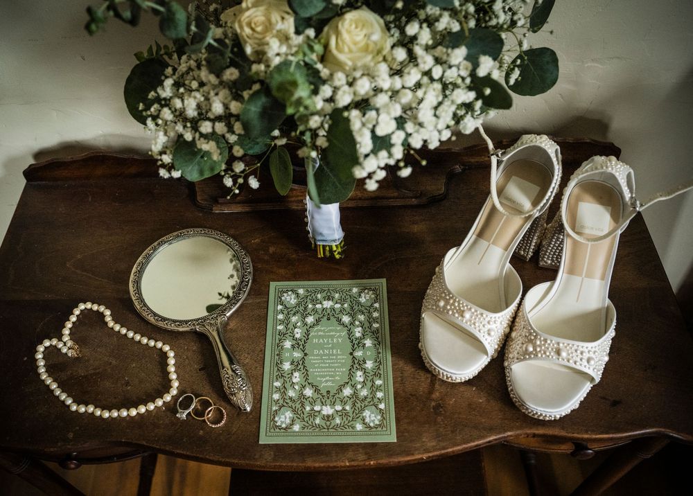 Elegant wedding accessories including white sandals, pearl necklace, mirror and floral arrangement on wooden table.