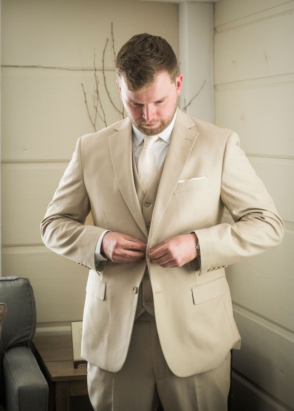 A groom in a light beige three-piece suit adjusts his jacket buttons while getting ready in a room.