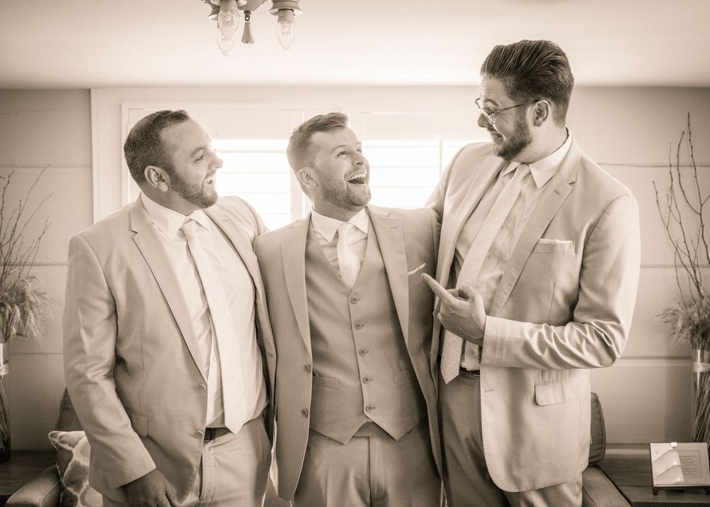 Three groomsmen in light colored suits share a candid laughing moment at a wedding in sepia tones.