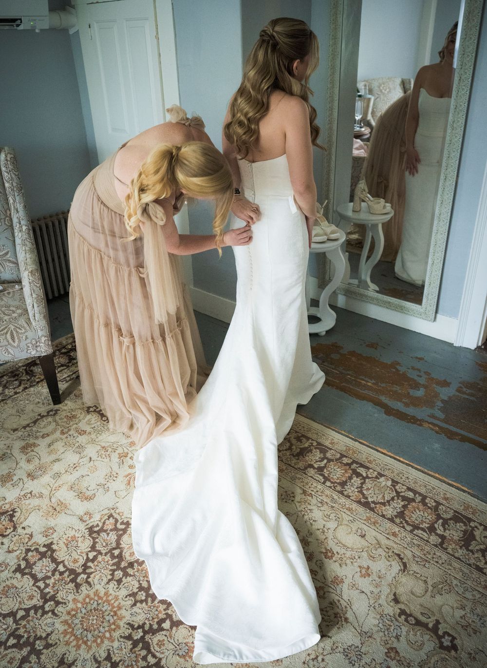 A person in a white wedding gown being helped with their dress by an attendant in a blush pink dress.