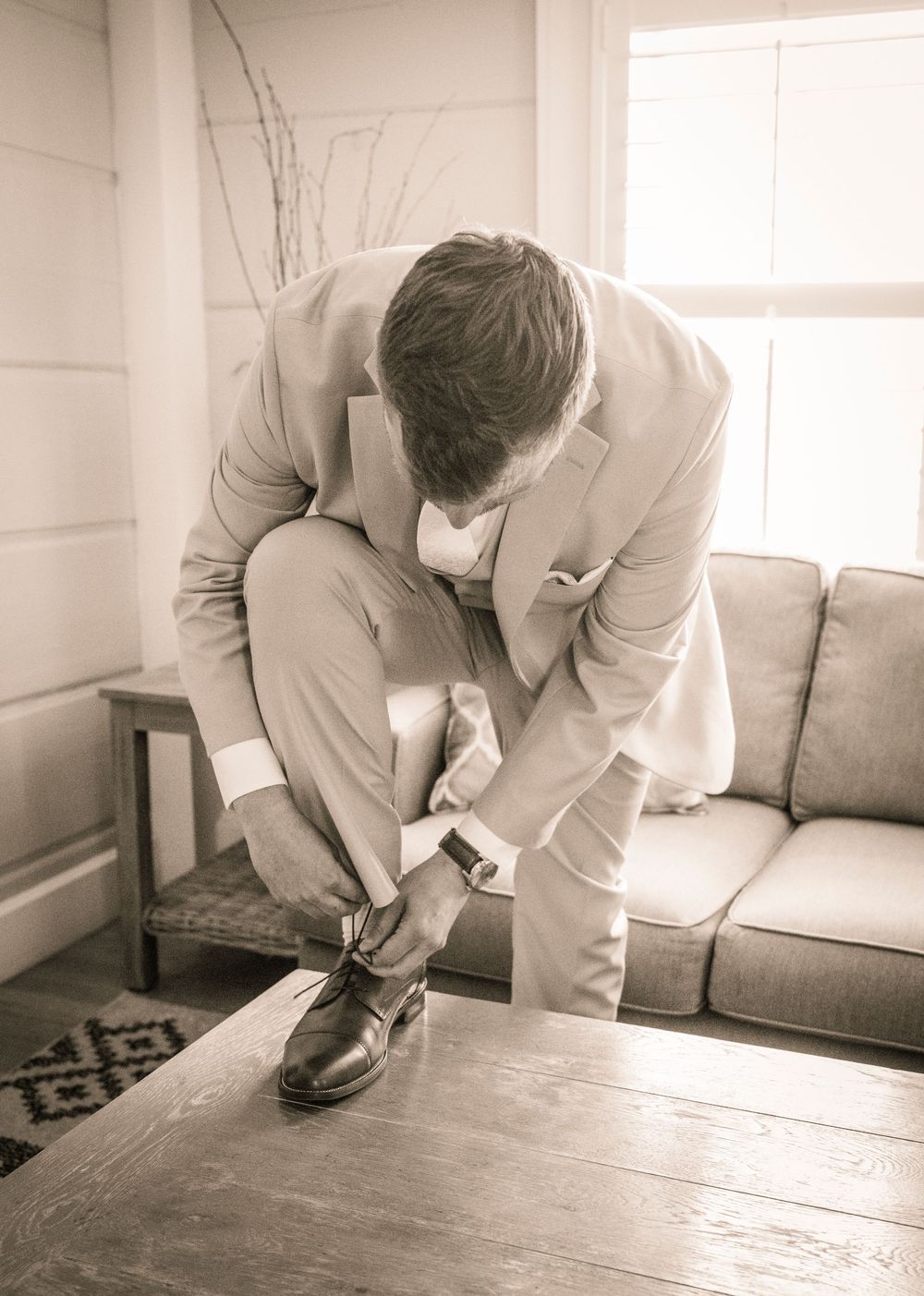 Figure in a light-colored suit bends down to tie dress shoes in a minimalist room with a grey couch and patterned rug.