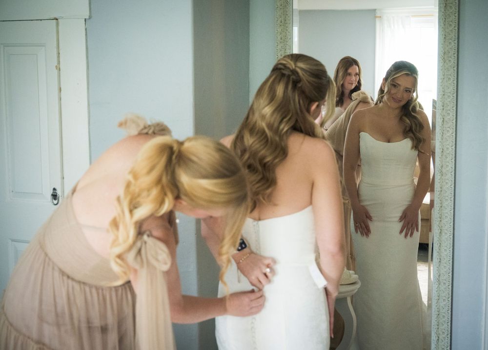 Friends help a bride with final adjustments to her wedding dress while looking in a mirror.