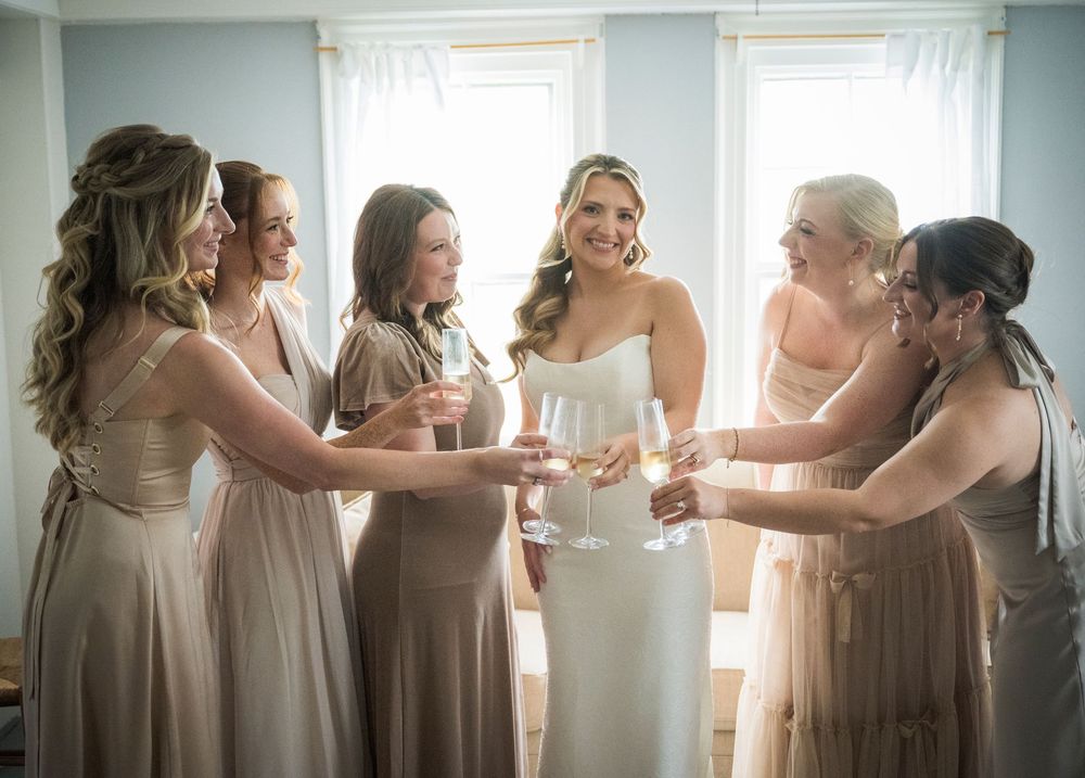 A group in champagne-colored bridesmaid dresses gather by sunlit windows to share a toast before the wedding ceremony.