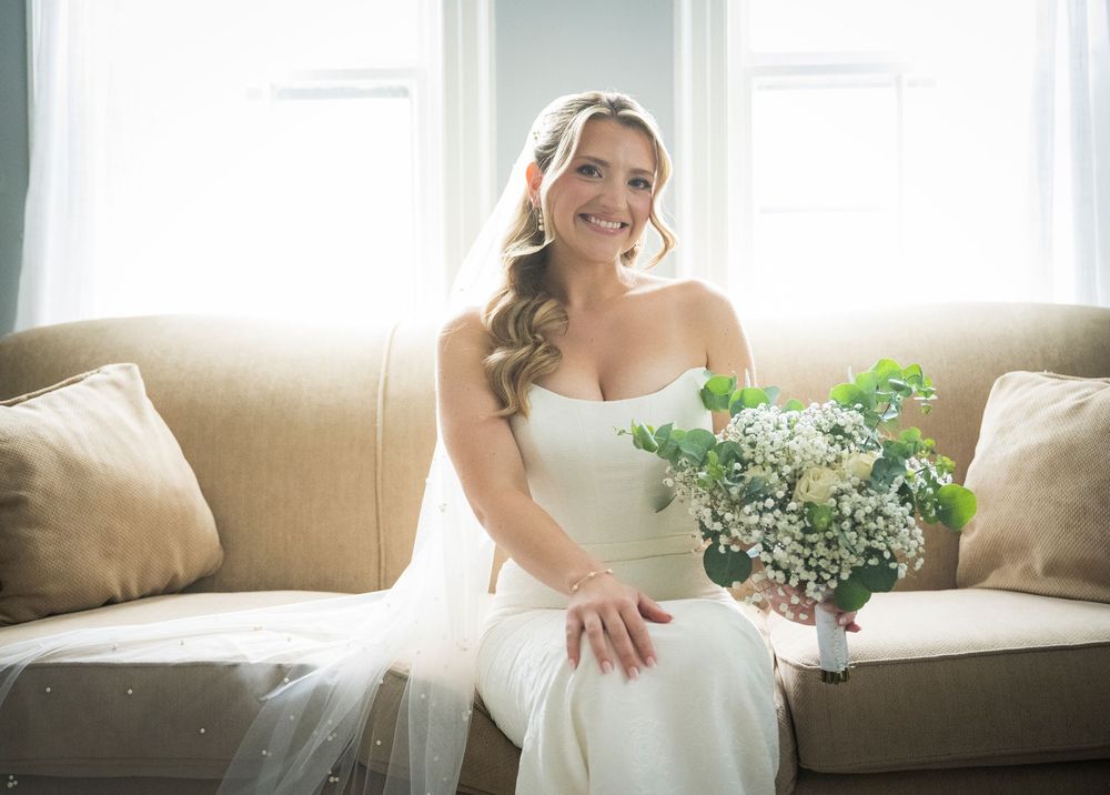 A bride in a white gown sits on a beige couch holding a bouquet of white and green flowers by sunlit windows.