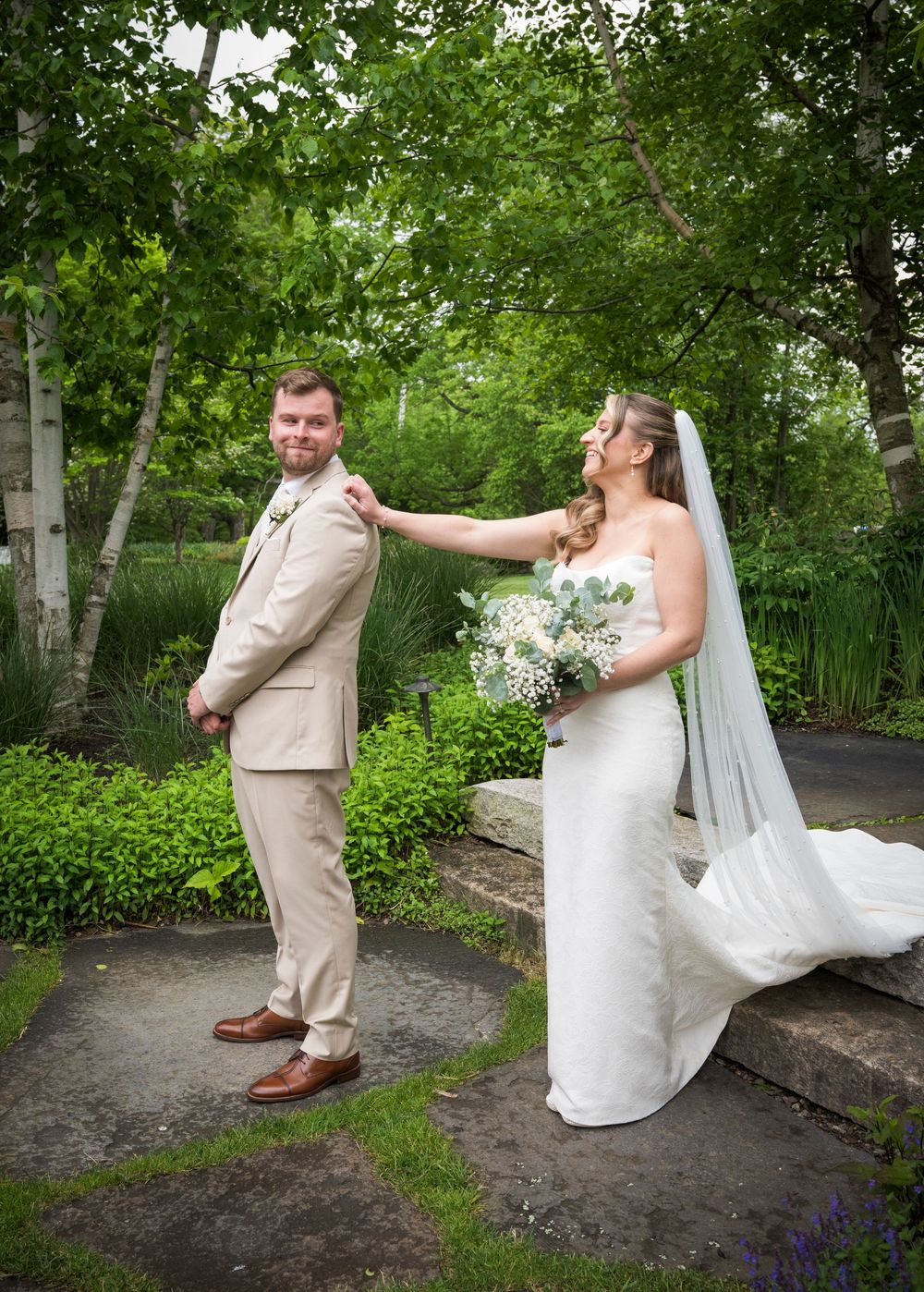 A couple shares a playful first look moment in a lush garden setting, with the bride in white and groom in beige.