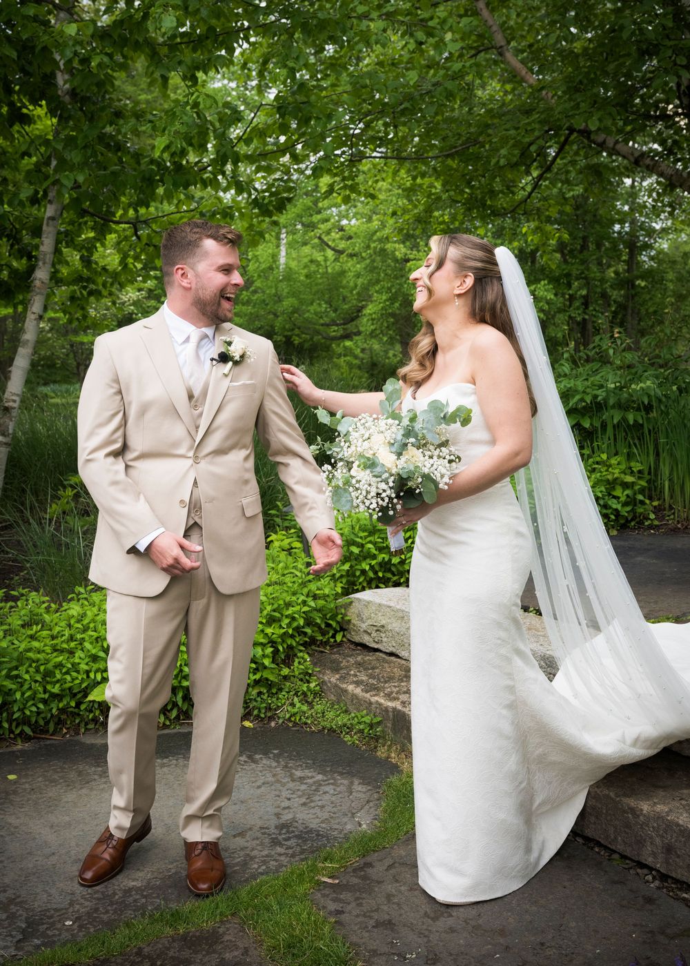 A couple shares a joyful moment together outdoors in formal wedding attire with lush greenery in the background.