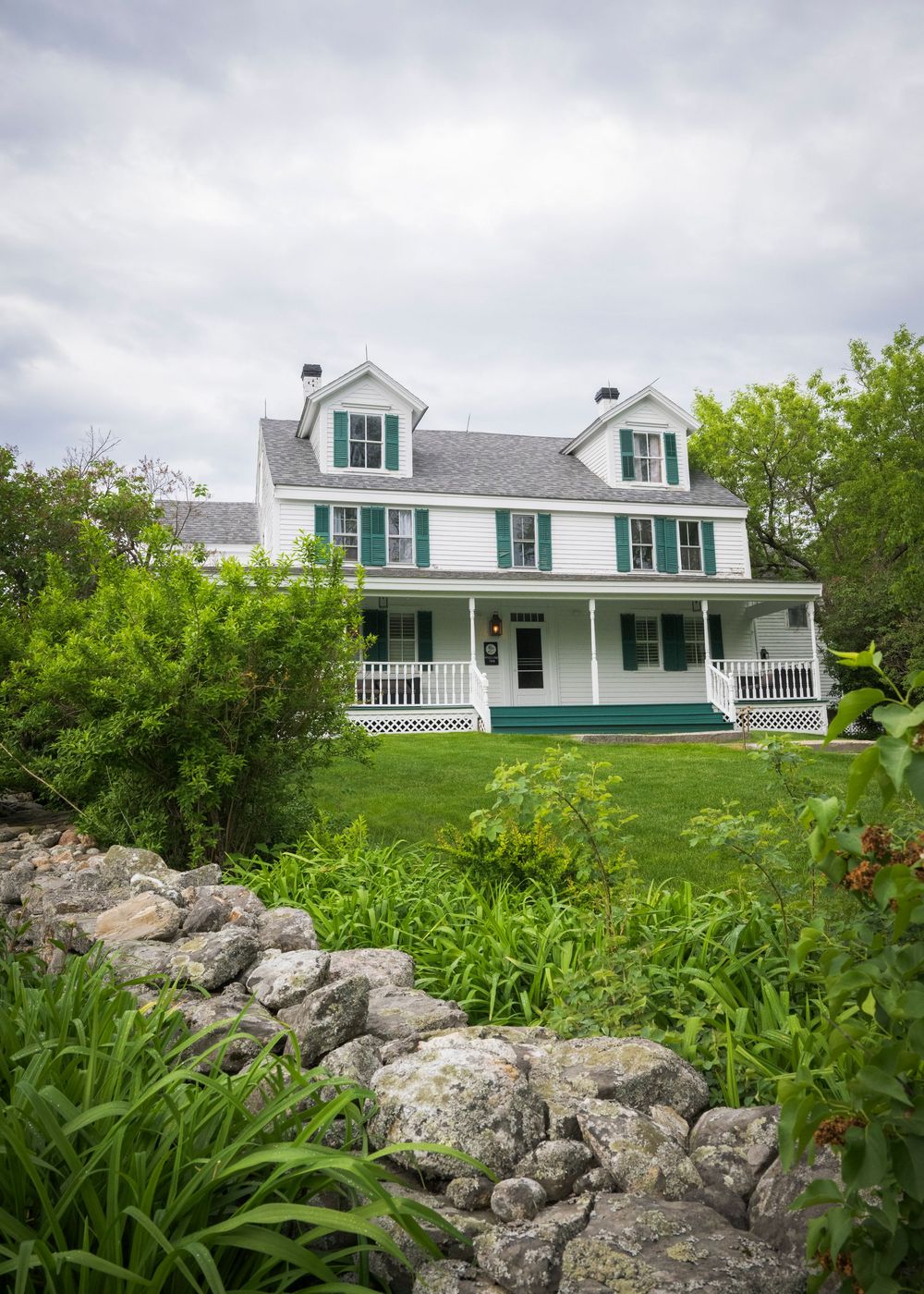 White colonial farmhouse with green shutters and wraparound porch sits atop landscaped yard with rustic stone wall.