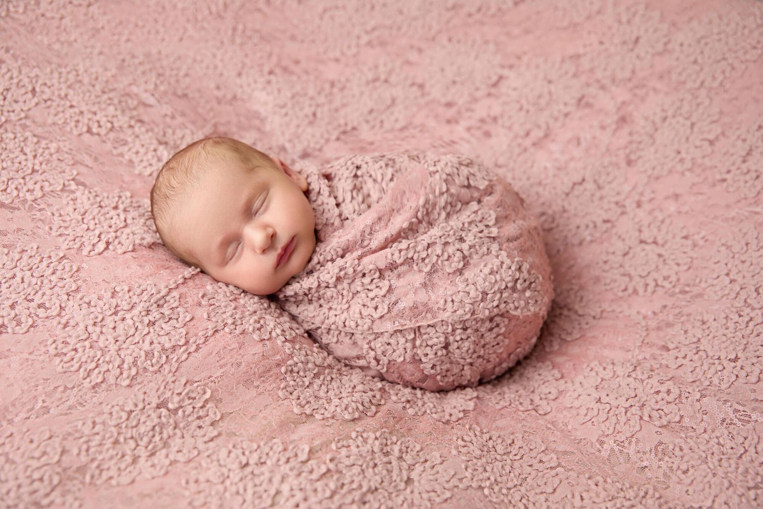 Sleeping newborn wrapped in soft pink textured blanket lying on matching pink backdrop for professional portrait.