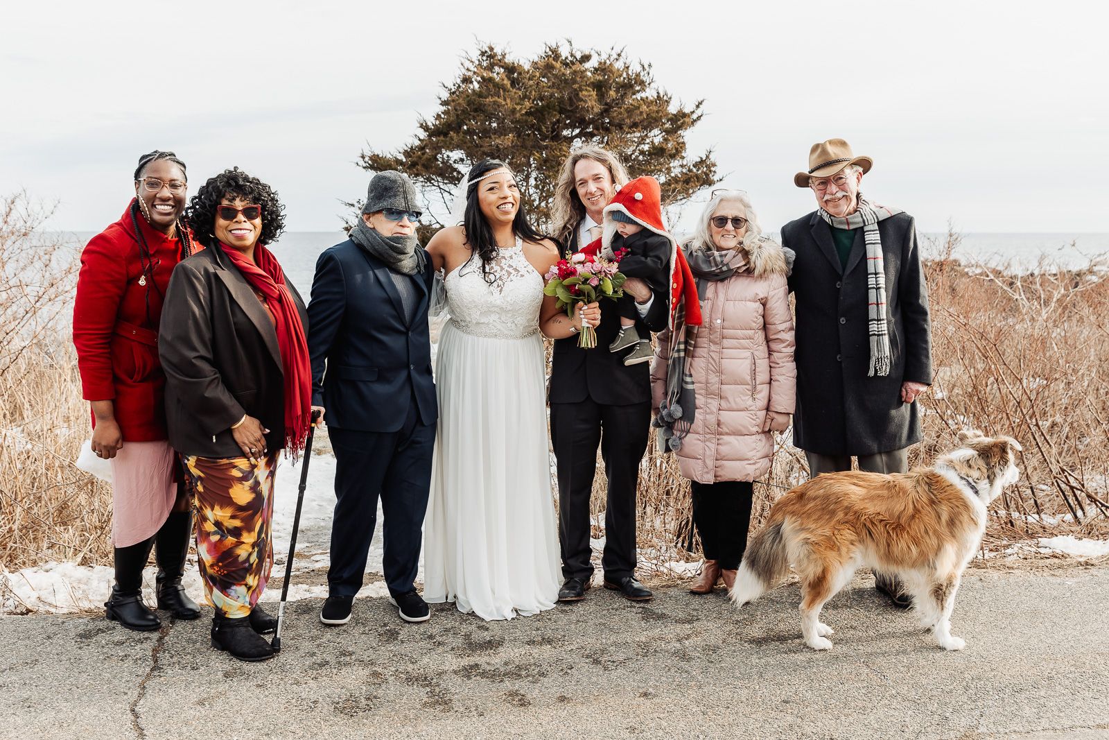 A group stands together outdoors in winter clothing with a bride in white and a fluffy dog on a leash.