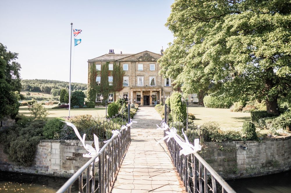 Stately brick mansion with white pillars and bridge entrance surrounded by lush greenery.