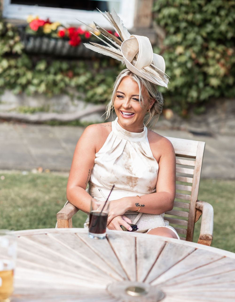 Wedding guest in elegant beige fascinator enjoying drinks at outdoor reception.