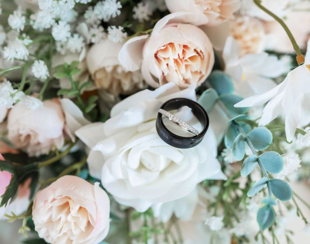 Black wedding band photographed among white and peach roses with eucalyptus leaves.