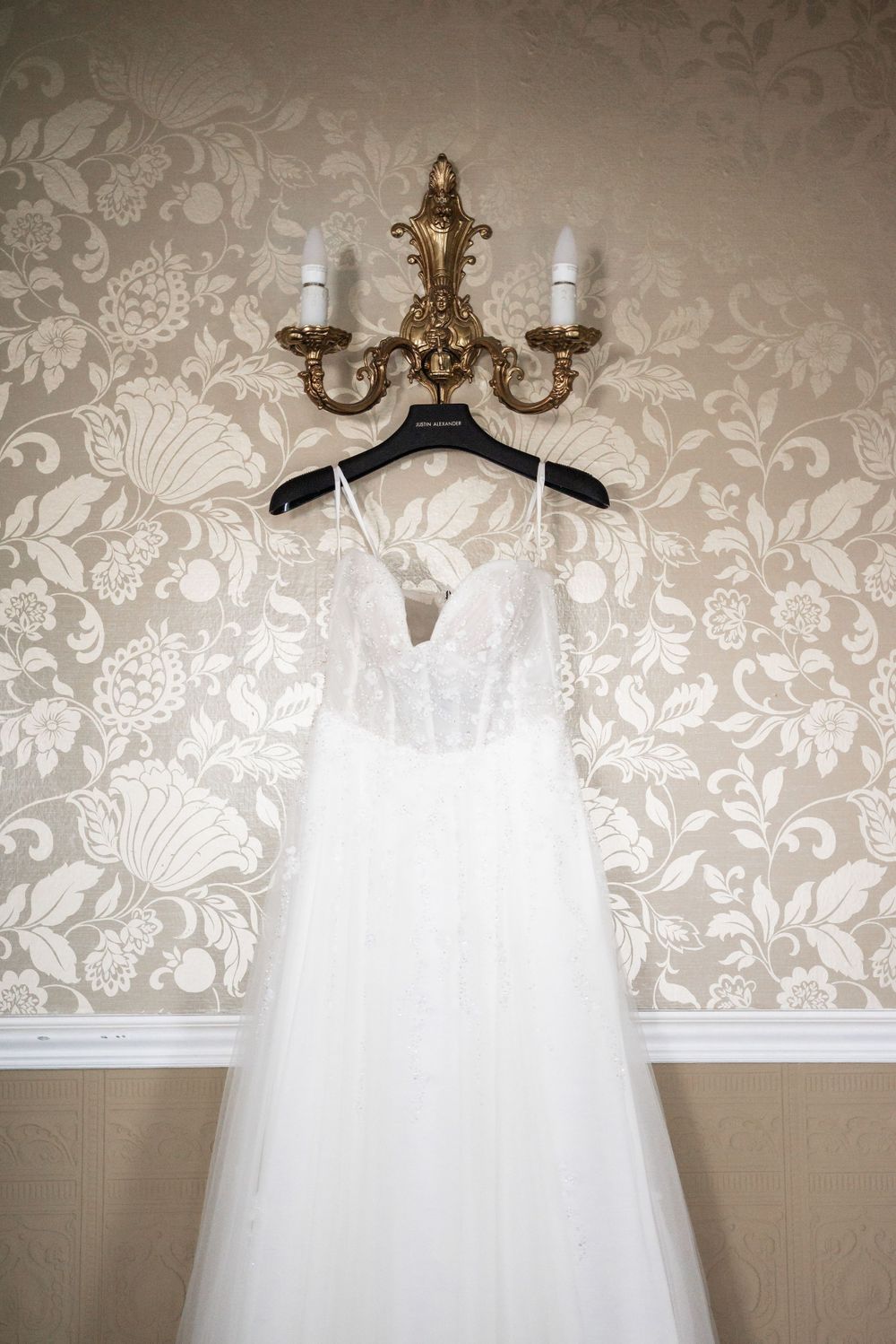 White wedding dress hanging on ornate gold chandelier against floral wallpaper.
