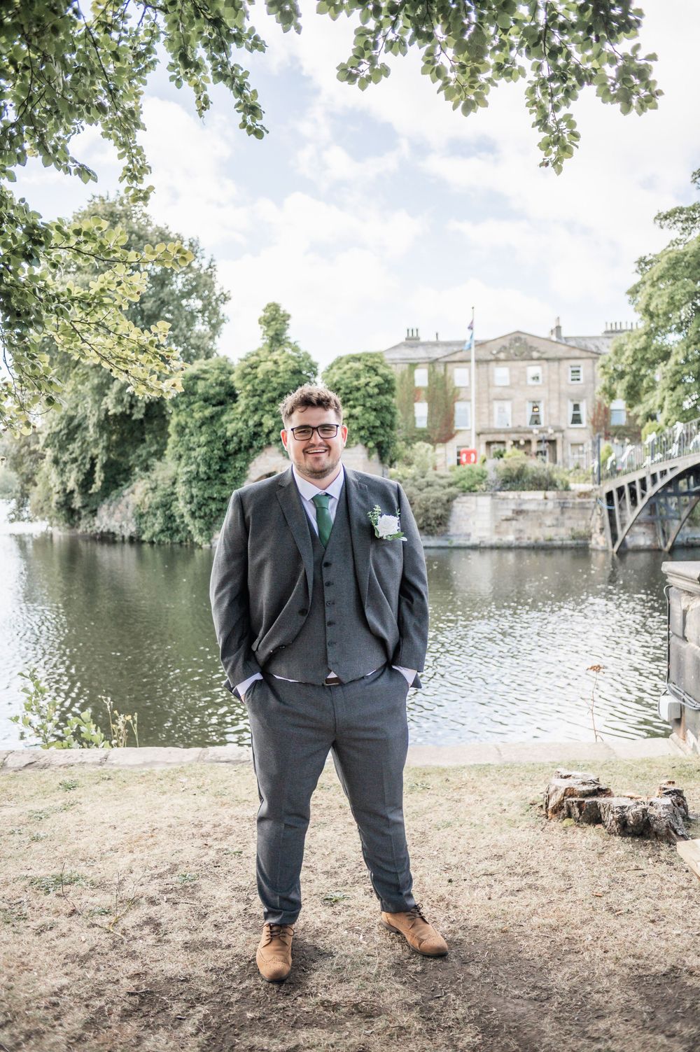 Wedding guest in grey suit standing by lakeside with historic building in background.