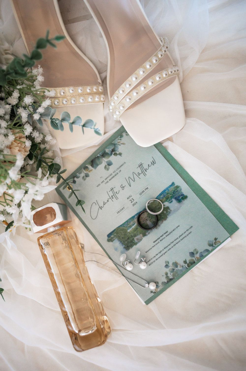Wedding details flat lay with pearl shoes, invitation, perfume and eucalyptus on white fabric.