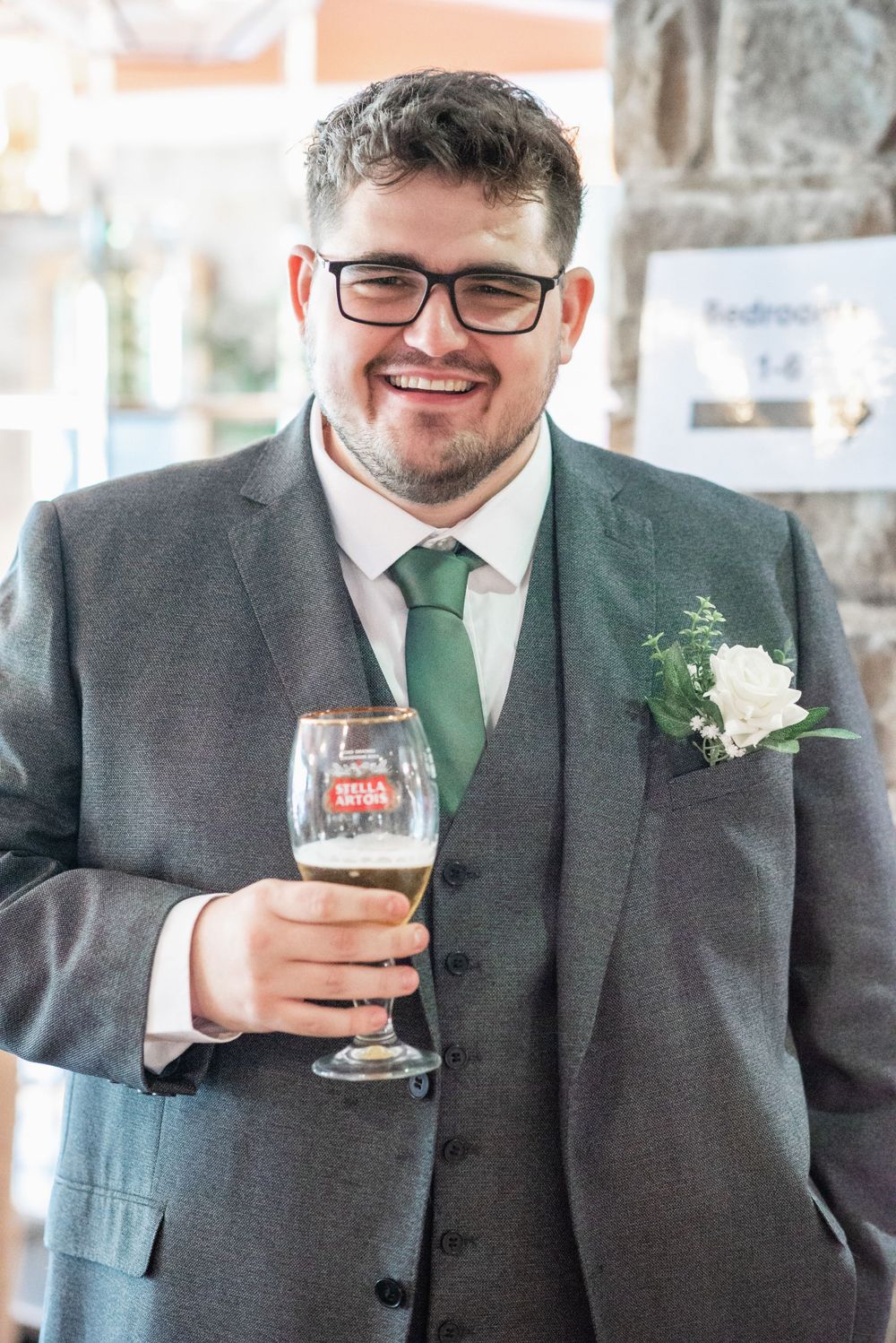 Wedding guest in grey suit and green tie holding beer glass at reception.