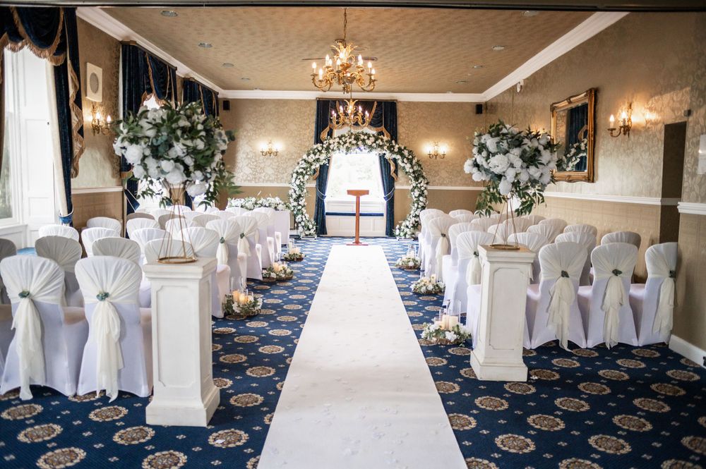 Elegant wedding venue setup with white floral arch, blue carpet, and white chair covers.